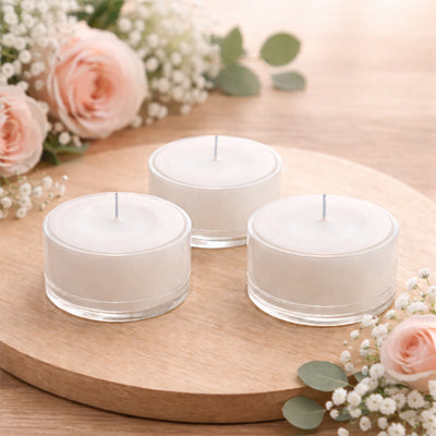 Three white tealight candles in glass holders on a wooden surface with flowers.