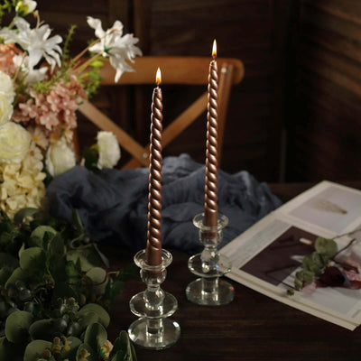 Twisted bronze candles in glass holders on a rustic table with floral arrangement.