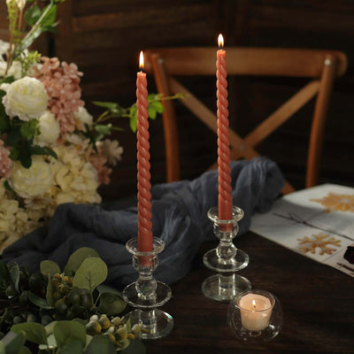 Twisted pink candles in glass holders on a rustic table with flowers.