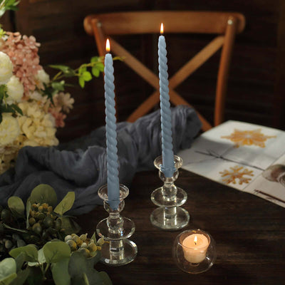 Blue twisted candles in glass holders on a rustic table with flowers and a candle.