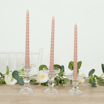 Twisted pink candles in glass holders with greenery backdrop.