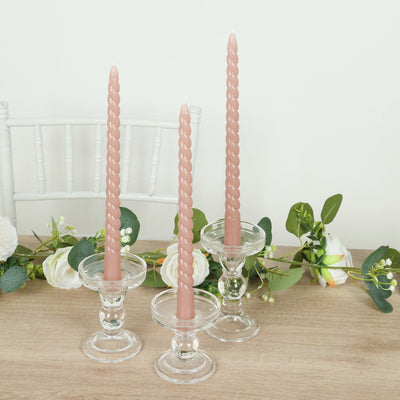 Twisted pink candles in glass holders with greenery backdrop.