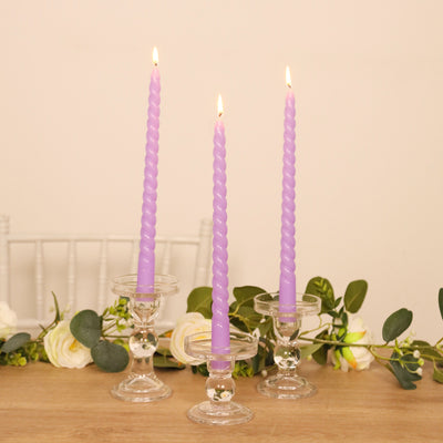 Three purple twisted candles in glass holders on a wooden table with flowers.