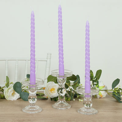 Purple twisted candles in glass holders with greenery backdrop.