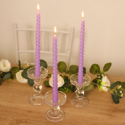 Purple twisted candles in glass holders on a wooden table with greenery