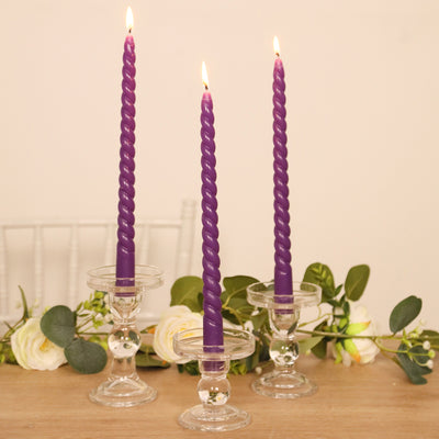 Three purple twisted candles in clear glass holders with greenery and roses.