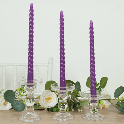 Purple twisted candles in clear glass holders on a table with flowers.