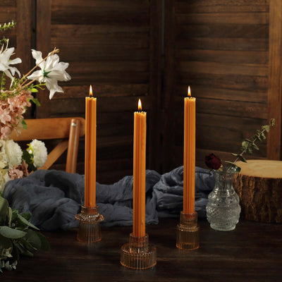 Three tall orange candles in clear holders on a wooden table with flowers.
