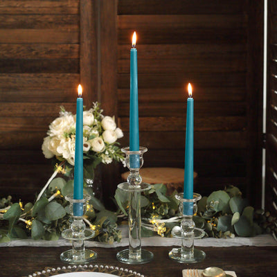 Three elegant teal taper candles in glass holders with flowers and greenery in the background.