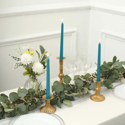 Teal candles in gold holders on a table with greenery and floral decor.