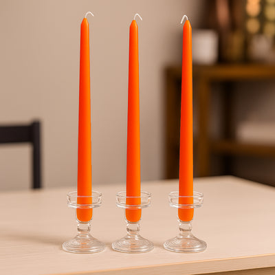 Three orange taper candles in clear glass holders on a table.