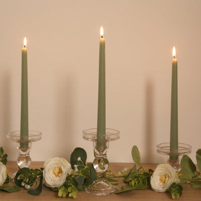 Green tapered candles in clear holders with floral accents on a wooden surface.