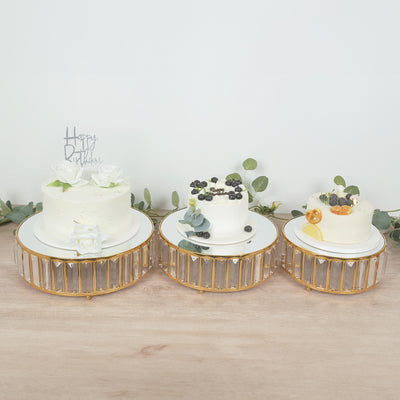 Three elegant birthday cakes on stylish pedestals with decorative flowers and fruits.