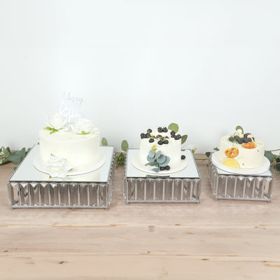Three elegantly decorated cakes on mirrored stands with greenery accents.