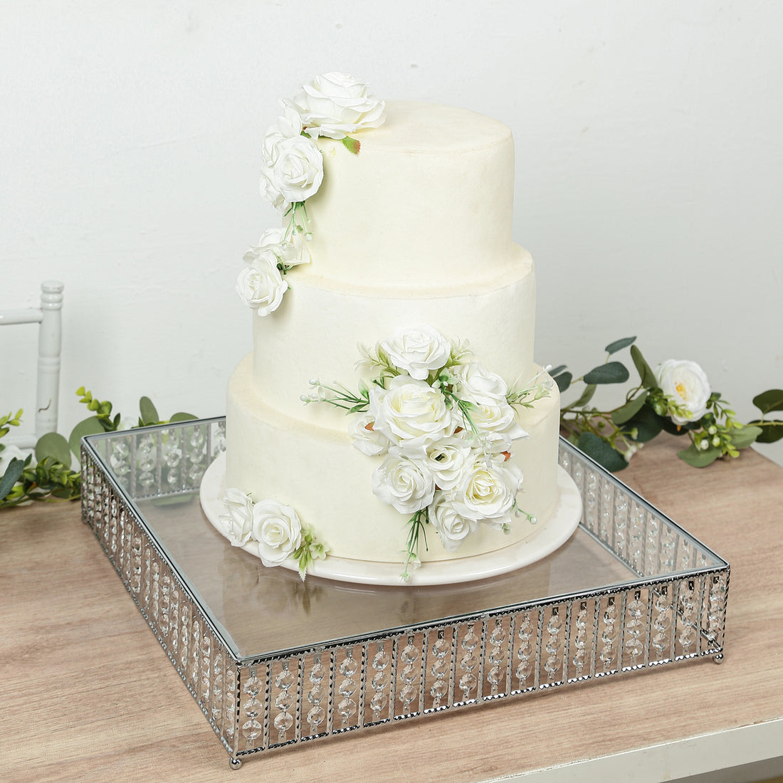 Elegant three-tier wedding cake adorned with white roses on a mirrored tray.