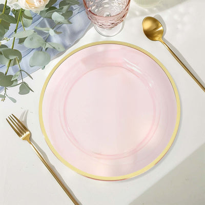 Pink plate with gold rim on a table setting with cutlery and greenery.