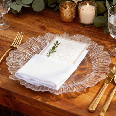 Elegant clear glass charger plate with a folded napkin and menu on a rustic wooden table.
