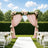 Outdoor wedding arch with pink drapes, flowers, and white chairs on a sunny lawn