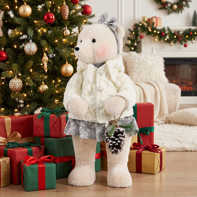 Christmas teddy bear doll in winter outfit beside decorated tree and gifts