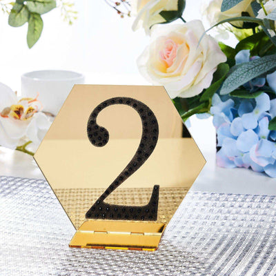 Gold hexagon table number 2 with black rhinestones on a textured table surface.