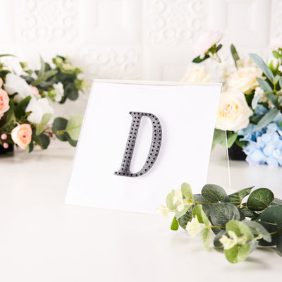 Decorative letter "D" with rhinestones on display with floral backdrop.
