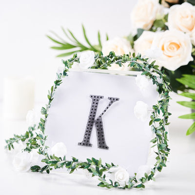 Decorative letter "K" framed with greenery and white flowers on a soft background.