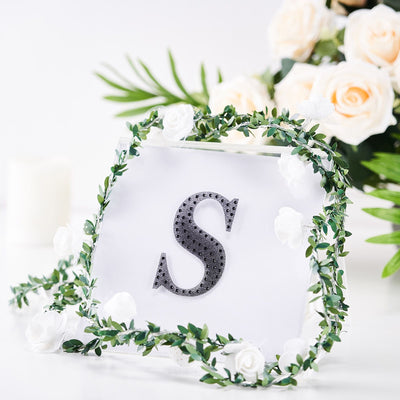 Black rhinestone letter "S" framed with greenery and flowers on a white background.