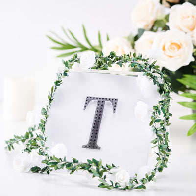 Decorative letter "T" framed with greenery and flowers on a clear backdrop.