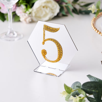 Gold glitter table number 5 in hexagon shape on a clear stand.