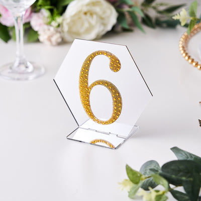Gold glitter table number 6 on a clear stand with floral decor in the background.
