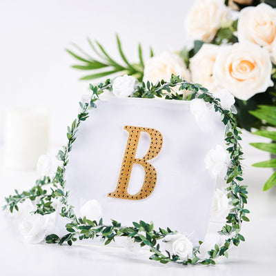Gold letter "B" adorned with greenery and white flowers on a clear backdrop.