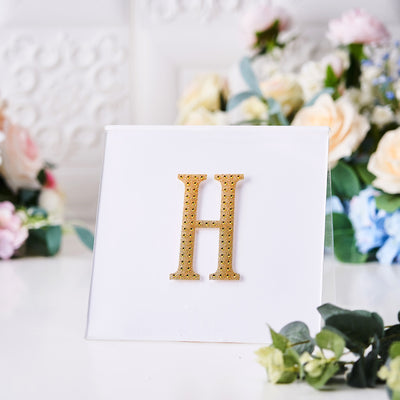 Gold glitter letter "H" on a clear display with floral background.