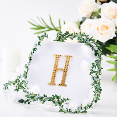 Gold letter "H" with floral greenery against a white backdrop.