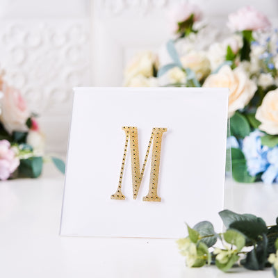 Gold rhinestone letter "M" decoration on a white card with floral background.