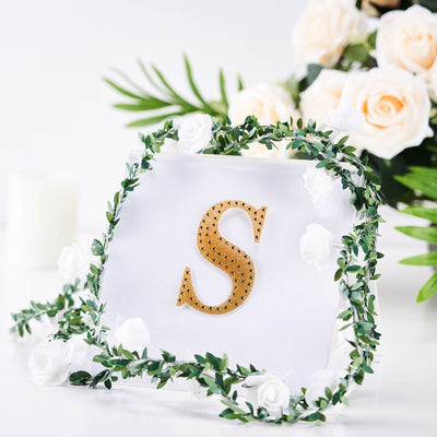 Gold letter "S" with greenery and white flowers on a plain background.