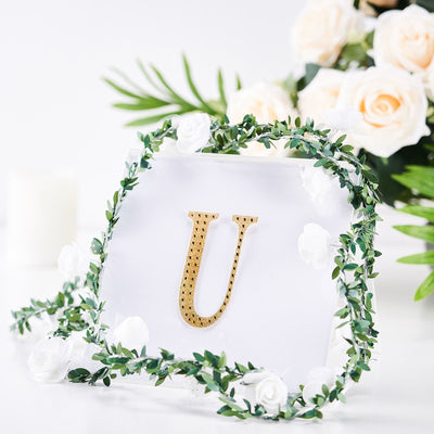 Gold letter "U" surrounded by greenery and flowers on a clear background.