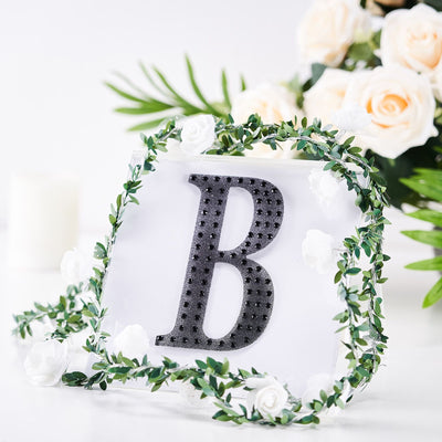 Black decorative letter "B" surrounded by greenery and white roses.