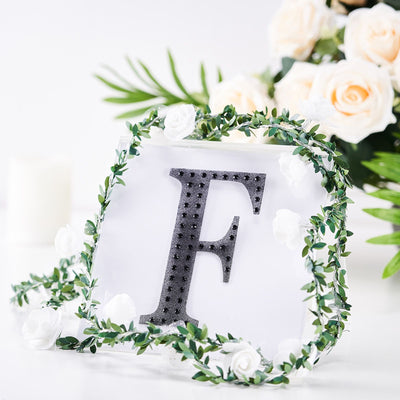 Decorative letter "F" surrounded by greenery and flowers on a white background.
