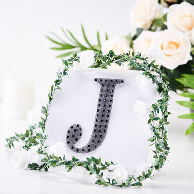 Black glitter letter "J" surrounded by greenery and white flowers.