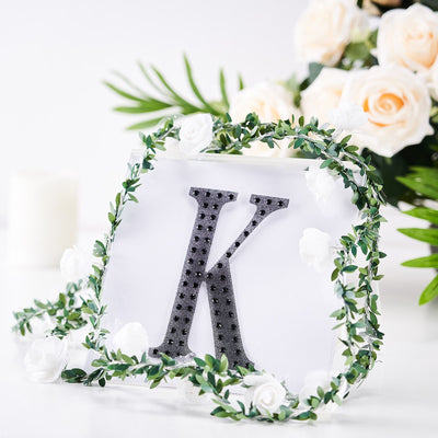 Black letter "K" adorned with greenery and white flowers on a white background.