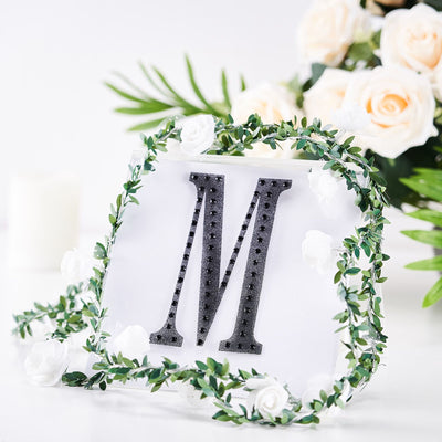 Decorative letter "M" surrounded by greenery and flowers in a bright setting.