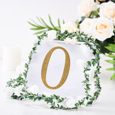 Gold number "0" sign adorned with greenery and white roses on a clear background.