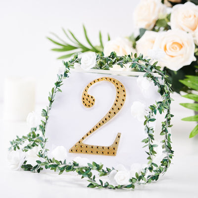 Gold glitter number 2 adorned with greenery and flowers on a white backdrop.