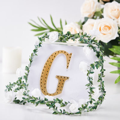 Gold letter "G" surrounded by greenery and flowers on a clear backdrop.