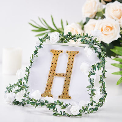 Gold letter "H" surrounded by greenery and white flowers on a white backdrop.
