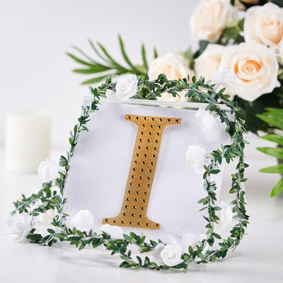 Gold letter "I" framed with greenery and white flowers on a soft background.