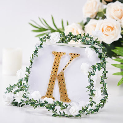 Gold letter "K" surrounded by greenery and white roses on a clear backdrop.