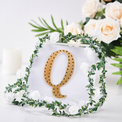 Gold letter "Q" decorative piece surrounded by flowers and greenery.