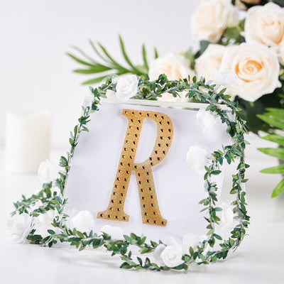 Gold letter R decorative sign with greenery and white flowers on a clear background.