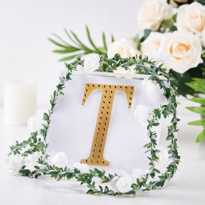 Gold letter "T" decorative sign surrounded by greenery and flowers.